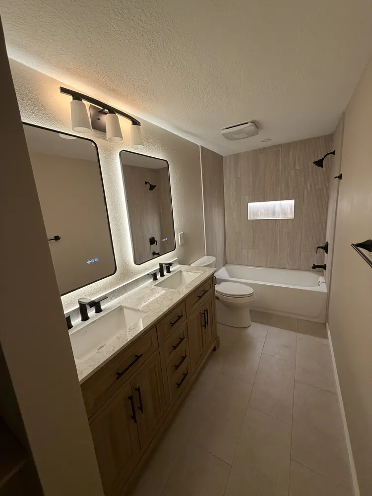 Remodeled bathroom with a wood double vanity, backlit mirrors, black fixtures and a wood-look tile tub surround