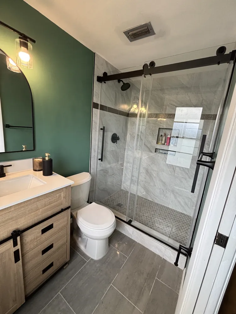 Bathroom with a deep green accent wall, light wood vanity, arched mirror and a barn-style glass shower door
