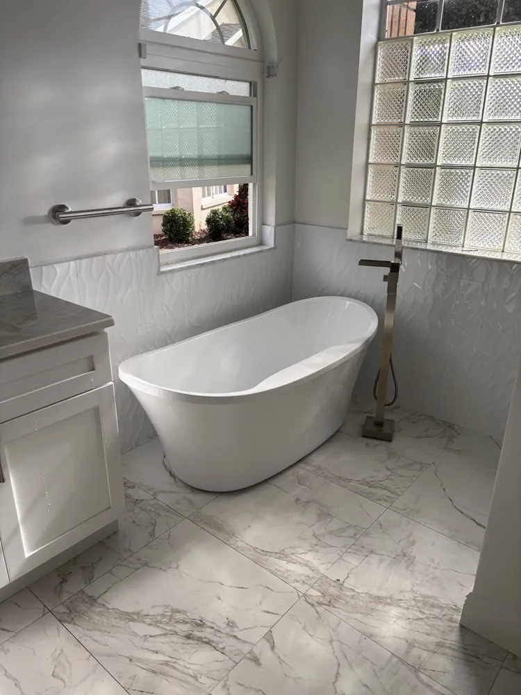 Freestanding tub beside a glass block window with a floor-mounted filler and marble-look floor
